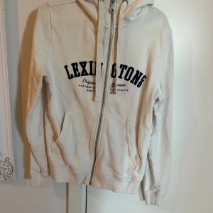 Vit hoodie från Lexington - Säljer en vit hoodie från Lexington med dragkedja och fickor framtill. Tröjan har svart tryck på bröstet och en huva med snören. Perfekt för en avslappnad stil och enkel att matcha med det mesta.