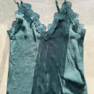 Mörkgrön ribbad linnetopp med spets - Snygg mörkgrön linnetopp med ribbad struktur och spetsdetaljer längs v-ringningen. Justerbara axelband för perfekt passform. Perfekt att styla till både jeans och kjol.