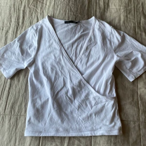 Vit omlottopp från Lager 157 - Säljer en vit omlottopp från Lager 157. T-shirten har korta ärmar och en snygg omlottdesign framtill. Perfekt till jeans eller kjol. I väldigt fint skick!