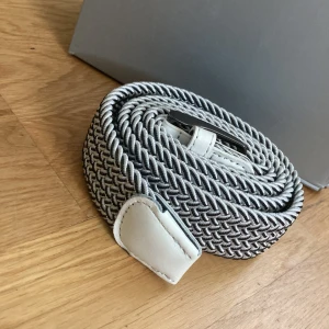 Vitt och grått flätat bälte - Snyggt flätat bälte i vitt och grått med silverfärgat spänne. Bältet har en cool, tvinnad design och detaljer i vitt skinn vid ändarna. Perfekt accessoar för att lyfta din outfit med en modern touch.