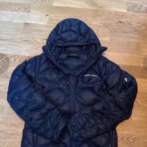 Mörkblå dunjacka från Peak Performance - Snygg mörkblå dunjacka från Peak Performance med huva och dragkedja framtill. Jackan är quiltad och har en sportig look, perfekt för kyliga dagar. Logga på bröstet och ärmen ger en clean vibe. Passar dig som gillar stilren och funktionell design.