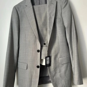 Ljusgrå kostym från Dressmann - Säljer en ljusgrå tvådelad kostym från Dressmann i slim fit-modell. Kavajen har två knappar, klassiska slag och fickor med lock. Byxorna har storlek 30/32. Perfekt för student eller sommarens tillställningar.