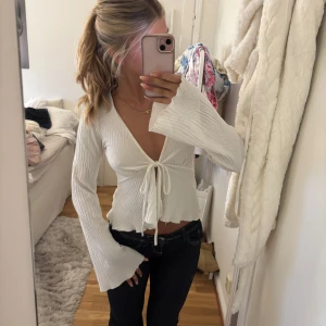 Vit ribbad omlottopp med knyt - Supersnygg vit omlottopp med djup v-ringning och långa trumpetärmar. Toppen är ribbad och har knyt framtill i midjan för en extra trendig look. Perfekt att styla med jeans eller kjol för en cool vibe.
