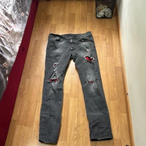 Grå jeans från Dsquared2 - Grå jeans från Dsquared2 med flera slitningar och hål, där röd tyg syns under vissa av hålen. Klassisk femficksmodell med raka ben och låg midja. Jeansen har en cool, edgy vibe och är tillverkade i bomull.