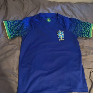 Blå Brasilien fotbollströja CBF - Snygg blå Brasilien fotbollströja med CBF-emblem broderat på bröstet. Ärmarna har leopardmönster i blått och grönt med gula detaljer vid mudden. Tröjan är kortärmad och tillverkad i ett lätt, ventilerande material som passar perfekt för match eller träning.