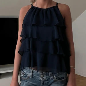 Mörkblå volangtopp med paljettdetalj - Säljer en mörkblå volangtopp med flera lager volanger och tunna axelband. Toppen har en glansig paljettkant upptill och är tillverkad i ett lätt, transparent material. Perfekt för sommarkvällar eller när du vill sticka ut lite extra.