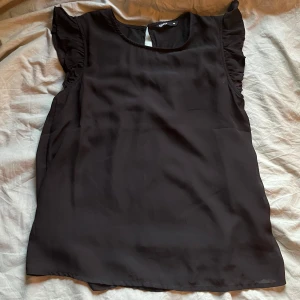 Svart blus med volangärmar - Säljer en svart blus med volangdetaljer vid ärmarna och rund halsringning. Blusen är ärmlös och har en knapp i nacken för enkel påklädning. Perfekt att styla till olika outfits.