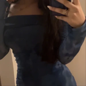 Blå långärmad kort tight klänning - Säljer en blå långärmad klänning med tight passform och offshoulder-detalj. Klänningen har ett diskret mönster i olika blå nyanser och sitter figurnära längs kroppen. Perfekt för dig som vill ha en stilren och trendig look.