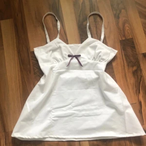 Vit topp med lila rosett🌷 - Söt vit topp med smala axelband och en dekorativ lila rosett framtill. Perfekt nu till sommaren då den är i 100% bomull 🌸