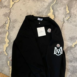 Svart sweatshirt från Moncler - Svart sweatshirt från Moncler med klassisk rund hals och långa ärmar. Tröjan har Moncler-logga på bröstet och ett stort vitt M-tryck med logga på ena ärmen. Tillverkad i mjuk bomull, perfekt för en clean och stilren look.  Strl S-M