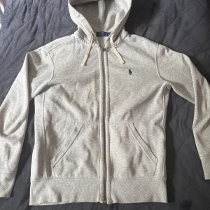 Grå hoodie från Polo Ralph Lauren - Snygg ljusgrå hoodie från Polo Ralph Lauren med dragkedja och huva. Klassisk broderad logga på bröstet och praktiska fickor framtill. Perfekt för chill dagar eller när du vill ha en clean och enkel stil.