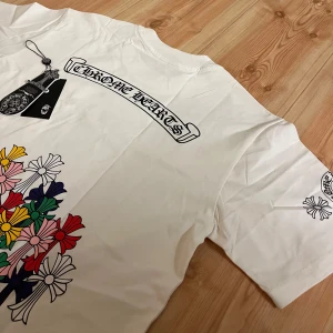 Vit Chrome Hearts t-shirt med blommor - Vit t-shirt från Chrome Hearts med coolt tryck av färgglada blommor på ryggen och logga på bröstet och ärmen. Klassisk passform och rund hals. Materialet är mjuk bomull, perfekt för en avslappnad streetwear-look.