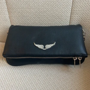 Svart Zadig axelväska med kedjedetalj - Snygg svart zadig axelväska i skinnimitation med silverfärgad kedjerem och glittrande vingdetalj framtill. Väskan har dragkedja både fram och bak, och en cool struktur i materialet. Perfekt för dig som vill ha en stilren men ändå unik accessoar.