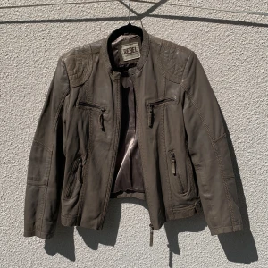 Brun/grå skinnjacka från REBEL - Snygg brun/grå skinnjacka från REBEL i äkta lammnappa. Jackan har flera dragkedjefickor framtill, quiltade detaljer på axlar och nedtill samt ståkrage. Knappt använd.