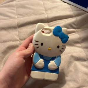 Hello Kitty blå mobilskal - Sött Hello Kitty mobilskal i form av figuren, med blå klänning och stor blå rosett. Skalet är gjort i mjukt syntetmaterial och har detaljer i vitt, blått och gult. Perfekt för dig som vill ha en gullig och unik look på mobilen. Den är för gammal telefonmodell