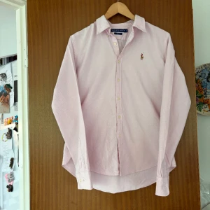 Ljusrosa skjorta från Ralph Lauren - Snygg ljusrosa skjorta från Ralph Lauren i slim fit-modell. Skjortan har klassisk krage, långa ärmar och knäppning framtill. Broderad logga på bröstet och tillverkad i mjuk bomull. Perfekt för dig som gillar stilrena och tidlösa plagg.