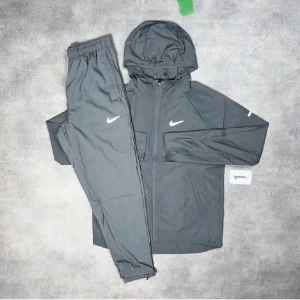 Grå Nike träningsset med huva - Snyggt grått träningsset från Nike med jacka som har huva och dragkedja samt matchande byxor. Båda plaggen har den klassiska vita Nike-loggan. Perfekt för dig som gillar sportig och clean stil.