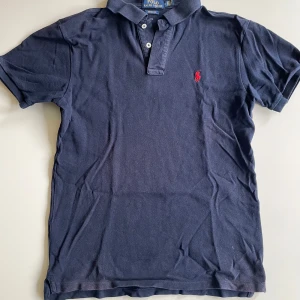 Marinblå pikétröja från Polo Ralph Lauren - Klassisk marinblå pikétröja från Polo Ralph Lauren med korta ärmar och krage. Tröjan har knäppning med tre vita knappar och den ikoniska röda loggan broderad på bröstet. Tillverkad i bomull. Säljer för 300kr