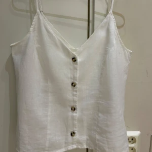 Vit linnetopp med knappar - Säljer en stilren vit linnetopp med smala axelband och knappar framtill. Toppen har v-ringning och är tillverkad i ett svalt linnematerial, perfekt för varma dagar.