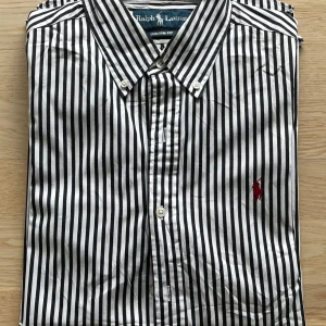 Randig skjorta från Ralph Lauren M - Snygg skjorta från Ralph Lauren i svart och vitt med vertikala ränder och röd broderad logga på bröstet. Modellen är custom fit med klassisk krage och knäppning framtill. Tillverkad i 100% bomull, perfekt för en clean och stilren look.