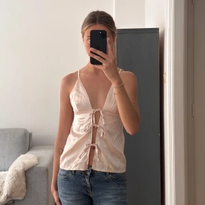 Ljusrosa satintopp med knytning - Säljer en ljusrosa topp i satin med smala axelband och djup v-ringning. Toppen har tre knytningar framtill. Perfekt för sommaren och ger en feminin look.🌸