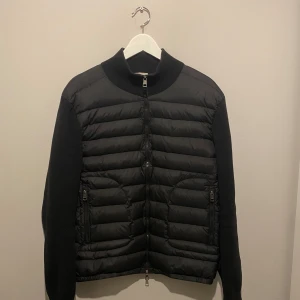 Svart Moncler cardigan  - Svart cardigan från Moncler med quiltad framsida och stickade ärmar. Jackan har hög krage, två fickor med dragkedja och Moncler-logga på ärmen. 