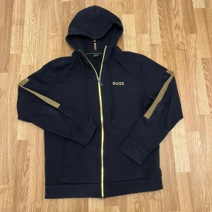 Svart zip tröja från BOSS - Svart hoodie från BOSS med guldfärgade detaljer på ärmarna och en guldfärgad dragkedja. Tröjan har huva med snörning och BOSS-logga på bröstet. Perfekt för dig som gillar stilrena och exklusiva streetwear-plagg. 