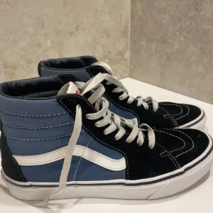 Vans skor - Säljer ett par klassiska Vans Sk8-Hi sneakers i blått och svart med vit sula och vit sidosnöre. Skorna har högt skaft, vit snörning och den ikoniska vågiga Vans-loggan på sidan. Perfekta för en streetwear-look.