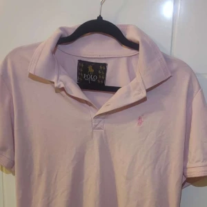 Ljusrosa pikétröja från Polo Ralph Lauren - Snygg ljusrosa pikétröja från Polo Ralph Lauren med klassisk krage och korta ärmar. Tröjan har den ikoniska broderade loggan på bröstet och är gjord i mjuk bomull. Perfekt för en chill och stilren look.