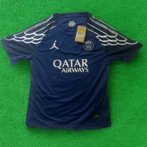 PSG fotbollströja blå Jordan x Paris Saint-Germain - Mörkblå PSG fotbollströja med vita detaljer och vingmönster på axlarna. Framsidan har Paris Saint-Germain-logga, Jordan-logga och Qatar Airways-tryck. Kortärmad och tillverkad i lätt, sportigt material.