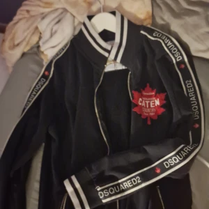 Svart bomberjacka från DSQUARED2 - Svart bomberjacka med vita detaljer och DSQUARED2-tryck längs ärmarna. Röd lönnlöv-patch med text på bröstet. Jackan har dragkedja och ribbade muddar vid krage, ärmslut och nederkant.