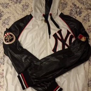 New York Yankees vindjacka från Majestic - Säljer en snygg New York Yankees vindjacka från Majestic. Jackan är vit med svarta ärmar, röda detaljer och broderade Yankees-loggor på bröstet och ärmen. Dragkedja framtill, huva med snörning och ribbade muddar.