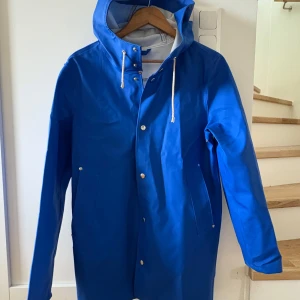 Stutterheim regn jacka - Säljer en blå regnjacka med huva och vita snören. Jackan har tryckknappar framtill och två fickor på sidorna. Perfekt för regniga dagar och enkel att matcha med andra plagg. Ok condition men finns fläckar (se bilder) men inte alla syns , kan skicka mera bilder