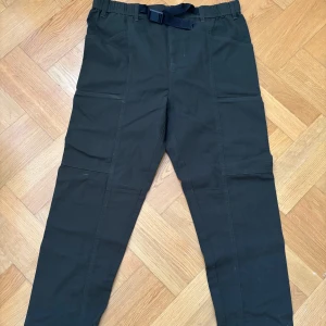 Mörkgröna cargobyxor från Knowledge Cotton Apparel - Mörkgröna cargobyxor från Knowledge Cotton Apparel med elastisk midja och inbyggt bälte. Byxorna har flera fickor och raka ben, perfekta för en avslappnad stil. Tillverkade i bomull för skön komfort.