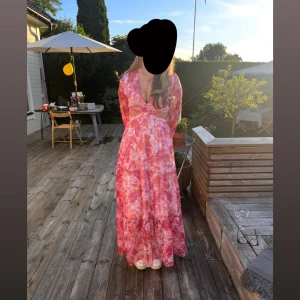 Rosa blommig långklänning med omlott och volang - Säljer en romantisk långklänning i rosa toner med blommigt mönster. Klänningen har omlottmodell, v-ringning och långa ärmar. Volangdetaljer och öppen rygg ger en extra touch. Perfekt för sommarens tillfällen!