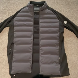 Svart hybridjacka från Sail Racing - Säljer en svart hybridjacka från Sail Racing i storlek small. Jackan har quiltade paneler fram och bak, dragkedja och två sidofickor med dragkedja. Perfekt för lager-på-lager och sportig stil.