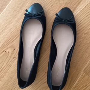 Svarta ballerinaskor med rosett från Mango - Säljer ett par klassiska svarta ballerinaskor från Mango med rund tå och liten rosett framtill. Skorna har en mörkgrå tådel i kontrast mot resten av skon och en platt sula. Perfekta för en stilren look. Inga defekter på ovansidan, endast lite på undersidan som inte syns när man har dom på sig🥰