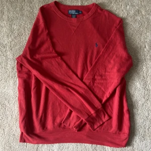 Röd tröja från Polo Ralph Lauren - Röd långärmad tröja från Polo Ralph Lauren med klassisk rund halsringning och broderad blå logga på bröstet. Tröjan är i mjuk bomull och har ribbade muddar vid ärmslut och nederkant. Perfekt för en stilren och avslappnad look.