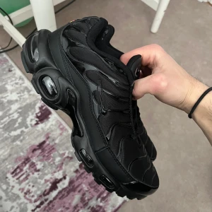 Svarta Nike Air Max Plus Tn - Säljer ett par helsvarta Nike Air Max Plus sneakers med ikonisk vågig design och synliga luftbubblor i sulan. Skorna har en sportig look med mesh och syntetdetaljer, samt rund tå och platt sula. Perfekta för dig som gillar streetwear och vill ha en clean stil.