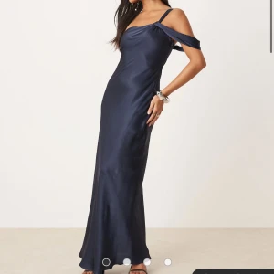 Marinblå långklänning med offshoulder - Snygg marinblå långklänning i satin med offshoulder och smala axelband. Klänningen har en elegant, figurnära passform och ett stilrent, slätt tyg som ger en lyxig känsla. Perfekt för speciella tillfällen.