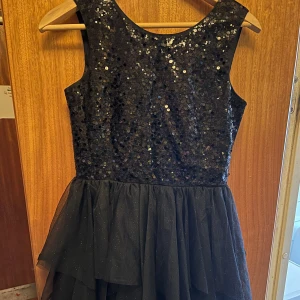 Svart glittrig klänning från H&M - Supersnygg svart klänning från H&M med paljetter upptill och flera lager tyll i kjolen. Klänningen har ärmlös design och rund halsringning. Perfekt för dig som vill sticka ut med lite extra glitter! (Barn)