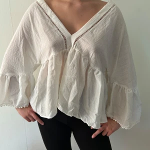 Vit blus med volangärmar och spetsdetaljer - Luftig vit blus med djup v-ringning fram och bak, volangärmar och spetsdetaljer längs ringningen. Blusen har en lös passform och är tillverkad i ett tunt, lätt material. Perfekt för varma dagar.