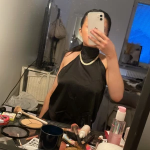 Svart halterneck topp i satin - Säljer en svart halterneck topp i glansigt satinmaterial. Toppen har hög hals och är ärmlös med öppen rygg, perfekt för en trendig look. Snygg att styla med smycken och passar till många olika outfits.