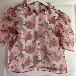 Blommig blus med puffärm från Pieces - Supersöt blommig blus från Pieces med volangkrage och puffiga korta ärmar. Topp i transparent, lätt material med rosa och beige rosor på vit botten. Perfekt för dig som vill sticka ut med romantisk vibe och trendig look.
