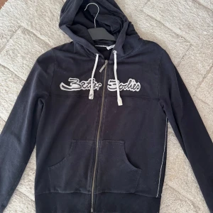 Svart hoodie med dragkedja från Better Bodies - Svart hoodie från Better Bodies med vit logga på bröstet och vita snören i huvan. Tröjan har dragkedja framtill och en stor ficka på magen. Den är Large men tycker ej att den är så stor 