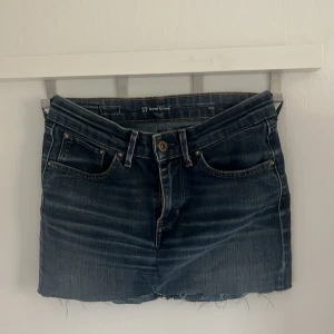 Mörkblå jeanskjol från Levi's - Säljer en mörkblå jeanskjol från Levi's med klassiska fickor både fram och bak. Kjolen har rå kant nertill och synliga sömmar i kontrastfärg. Perfekt för dig som gillar en enkel och tidlös stil.