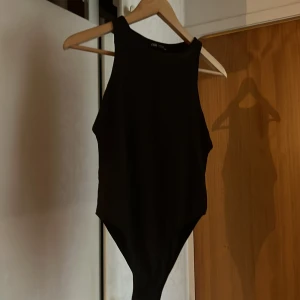 Svart ärmlös bodysuit från Zara - Säljer en svart, stilren bodysuit från Zara. Modellen är ärmlös med hög halsringning och har en tight passform. Perfekt att matcha med jeans eller kjol för en snygg look.
