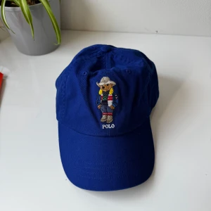 Blå keps från Polo Ralph Lauren med björn  - Snygg blå keps från Polo Ralph Lauren med broderad björn på framsidan. Justerbart spänne baktill i läder och klassisk Polo-logga broderad bak. Perfekt accessoar för en sportig och stilren look.