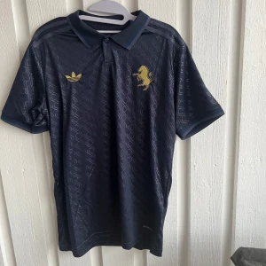 Svart Juventus fotbollströja med guldtryck - Snygg svart Juventus fotbollströja från Adidas med guldtryck och korta ärmar. Tröjan har ett diskret mönster och en klassisk krage. Perfekt för fotbollsfans som vill visa sitt stöd med stil. Nummer 10 och namnet Yildiz pryder baksidan.