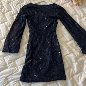 Mörkblå glittrig klänning från MEET x TIGER - Snygg mörkblå klänning från MEET x TIGER med glittriga paljetter över hela tyget. Klänningen har en v-ringad design i ryggen och långa ärmar, perfekt för en festlig kväll. Den är figurnära och ger en elegant look.